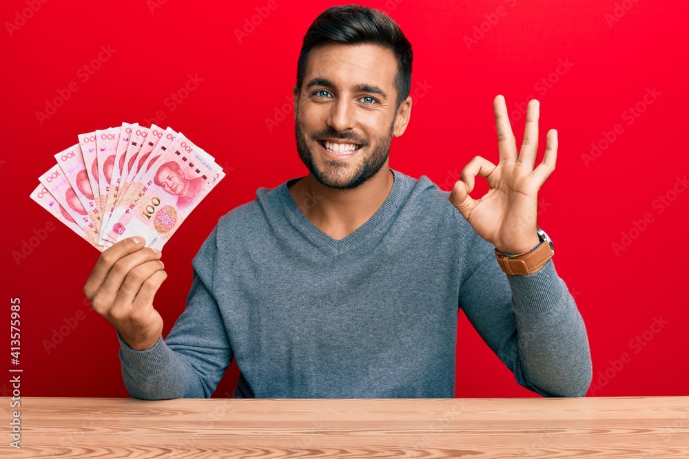 Handsome hispanic man holding yuan chinese banknotes doing ok sign with fingers, smiling friendly gesturing excellent symbol Handsome hispanic man holding yuan chinese banknotes doing ok sign with fingers, smiling friendly gesturing excellent symbol