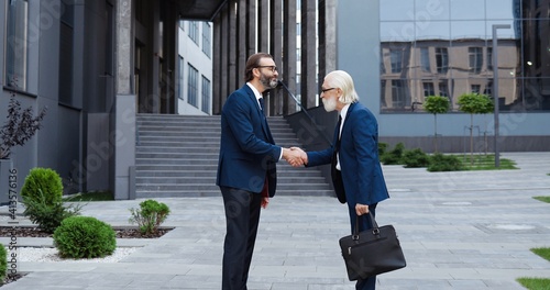 Caucasian senior businessmen in suits and ties meeting outdoor at business center, greeting with hands shaking and talking. Old rich men employees of office chatting. Business conversation in city. © VAKSMANV