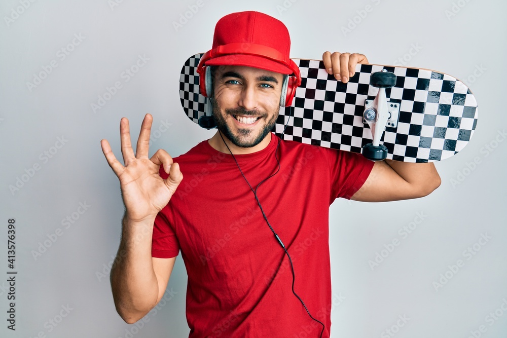 Young hispanic man using headphones holding skate doing ok sign with fingers, smiling friendly gesturing excellent symbol Young hispanic man using headphones holding skate doing ok sign with fingers, smiling friendly gesturing excellent symbol