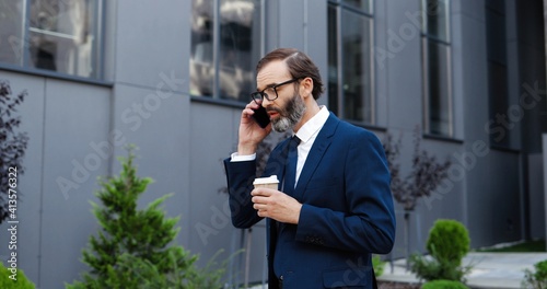 Photos Caucasian middle-aged man in glasses walking outdoor, talking on mobile phone and drinking coffe to-go in morning