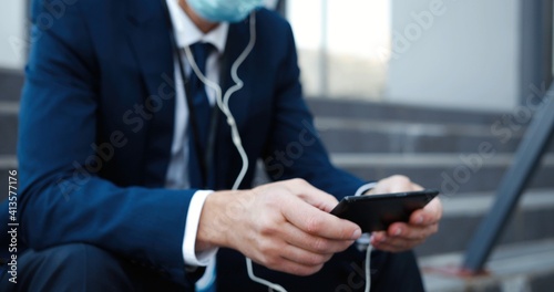 Caucasian middle-aged businessman in medical mask sitting on steps outdoor texting, tapping, playing or watching video on smartphone. Retired fired man in headphones listening to music on mobile phone