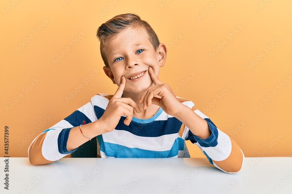 Adorable caucasian kid wearing casual clothes sitting on the table smiling with open mouth, fingers pointing and forcing cheerful smile Adorable caucasian kid wearing casual clothes sitting on the table smiling with open mouth, fingers pointing and forcing cheerful smile