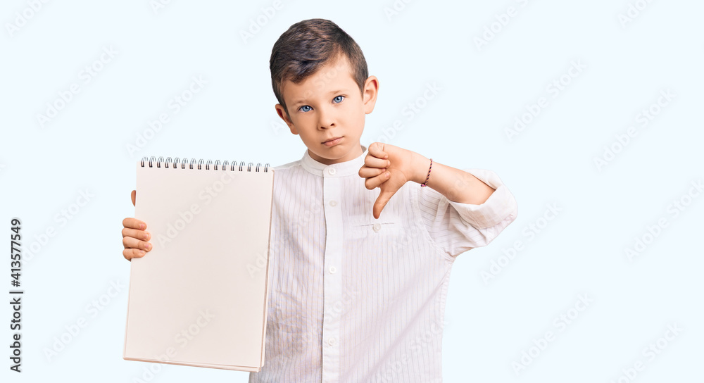 Cute blond kid holding notebook with angry face, negative sign showing dislike with thumbs down, rejection concept Cute blond kid holding notebook with angry face, negative sign showing dislike with thumbs down, rejection concept
