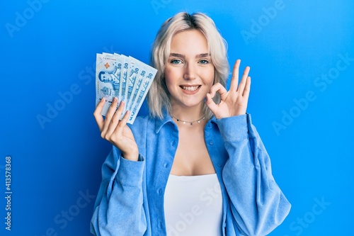 Photo Young blonde girl holding thai baht banknotes doing ok sign with fingers, smilin