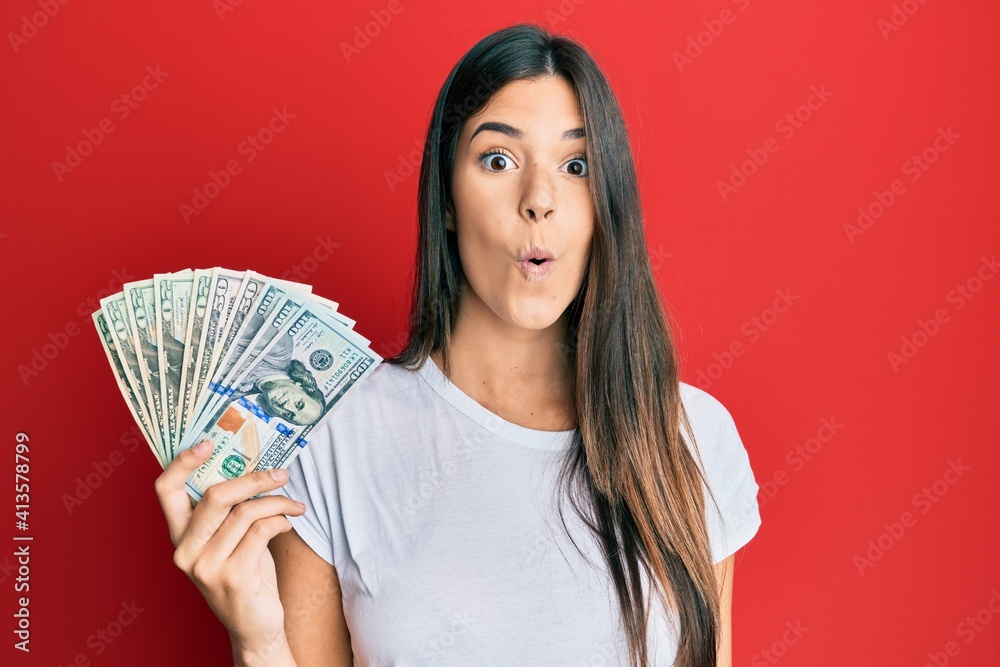 Young brunette woman holding dollars scared and amazed with open mouth for surprise, disbelief face Young brunette woman holding dollars scared and amazed with open mouth for surprise, disbelief face