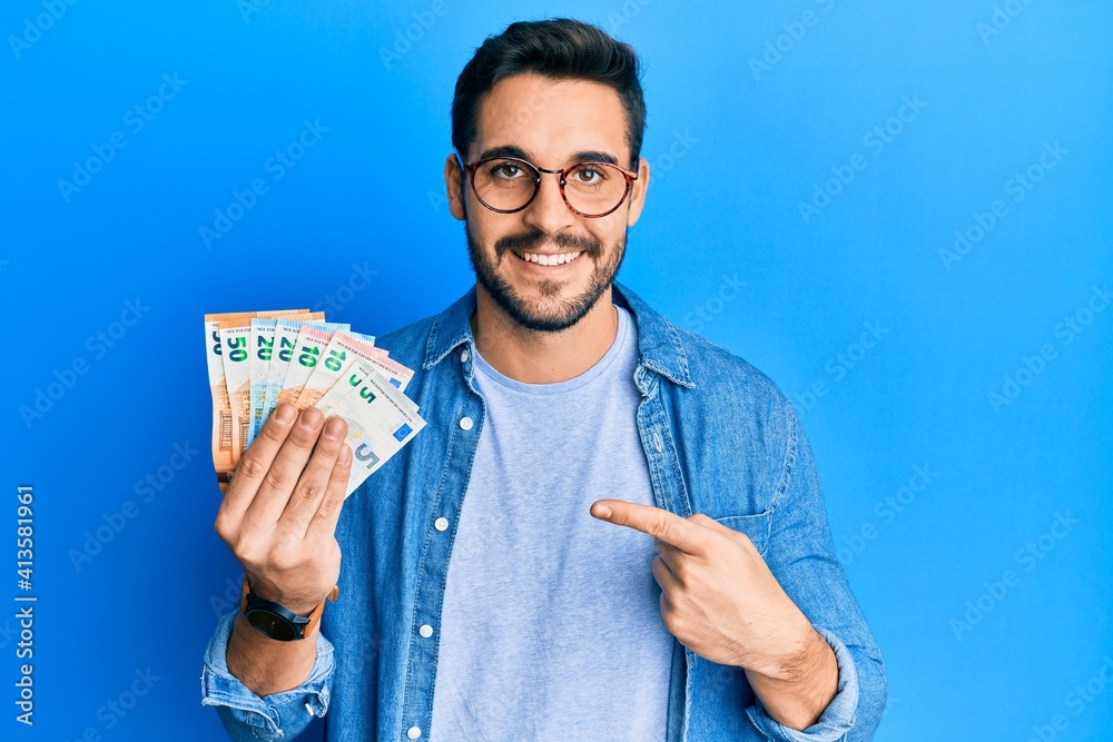 Young hispanic man holding euro banknotes smiling happy pointing with hand and finger Young hispanic man holding euro banknotes smiling happy pointing with hand and finger