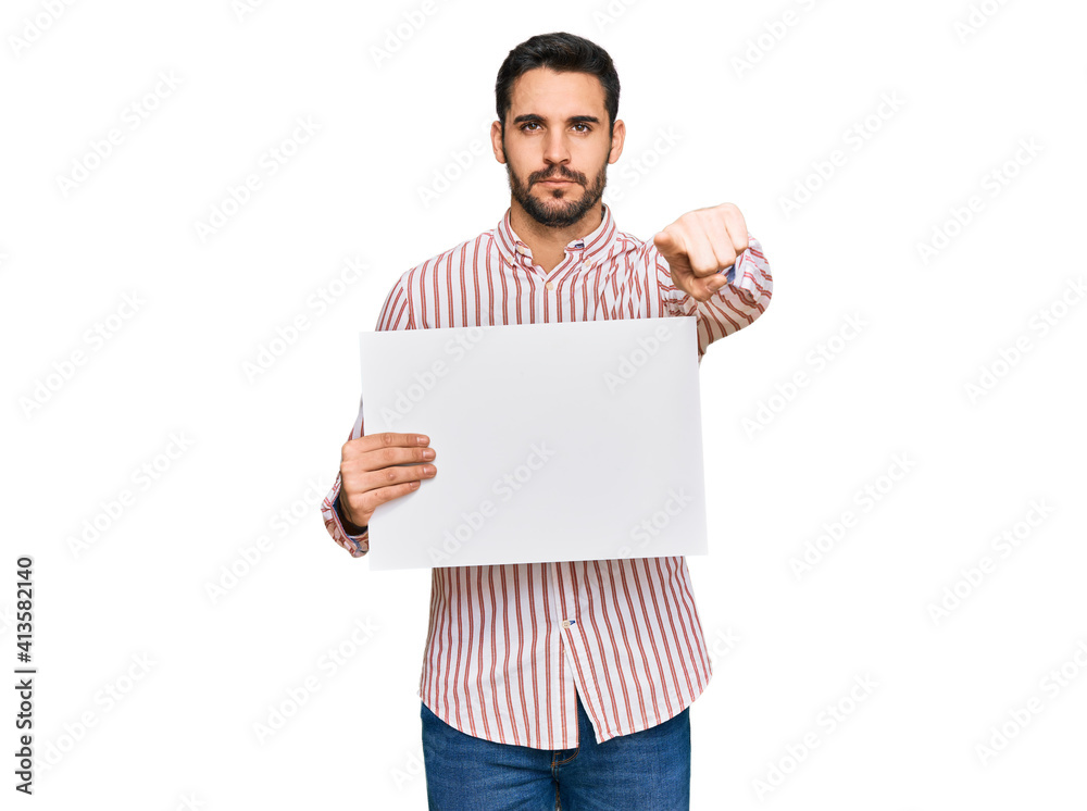 Young hispanic man holding blank empty banner pointing with finger to the camera and to you, confident gesture looking serious Young hispanic man holding blank empty banner pointing with finger to the camera and to you, confident gesture looking serious