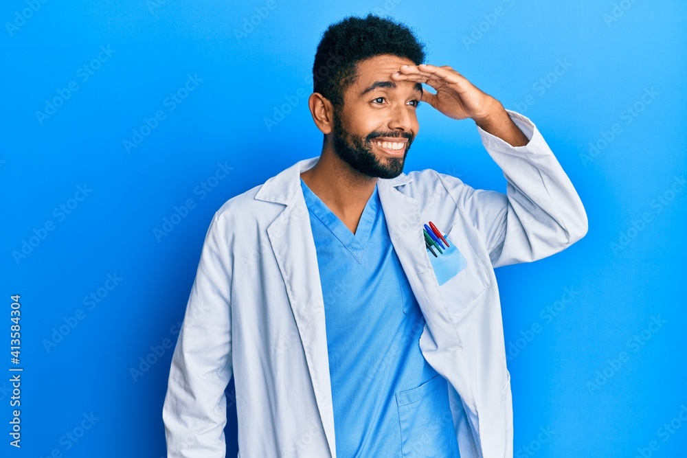 Handsome hispanic man with beard wearing doctor uniform very happy and smiling looking far away with hand over head. searching concept.