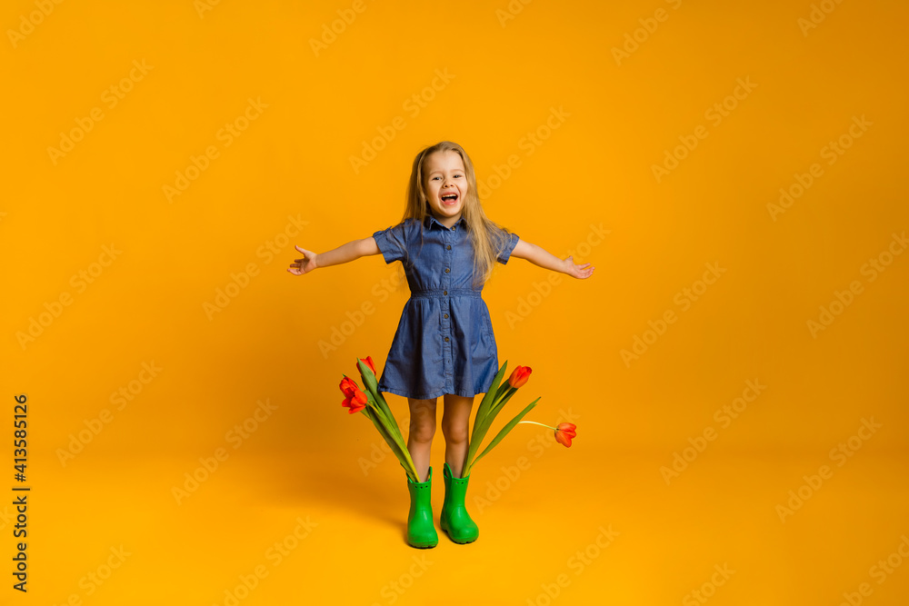 laughing little girl in green rubber boots with red tulips stands with her arms outstretched on a yellow background with space for text. mother's day