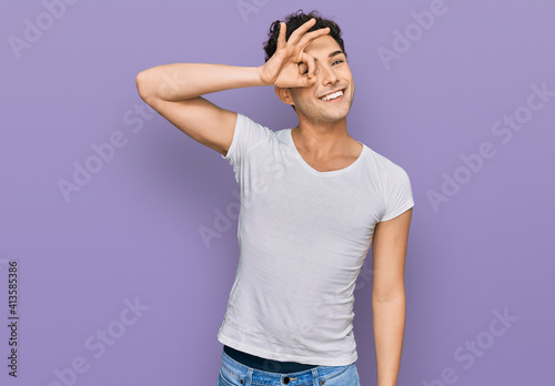 Young handsome man wearing casual white t shirt smiling happy doing ok sign with hand on eye looking through fingers