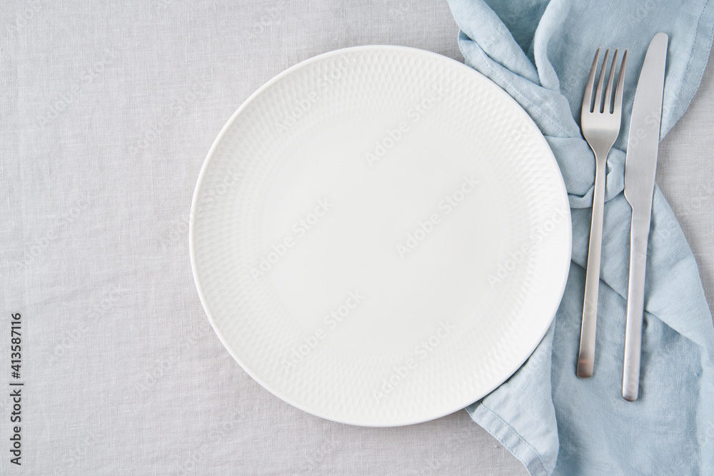 Clean empty white plate, fork and knife on pastel grey linen tablecloth on table, copy space, mock up, top view. Concept for menu with utensil