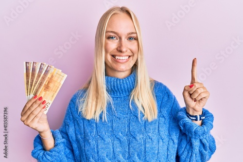 Young blonde girl holding 100 norwegian krone banknotes smiling with an idea or question pointing finger with happy face, number one photo