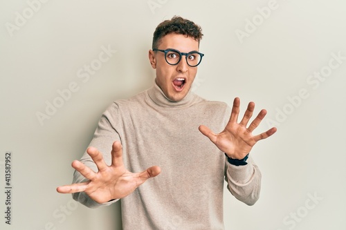 Hispanic young man wearing casual turtleneck sweater afraid and terrified with fear expression stop gesture with hands, shouting in shock. panic concept.