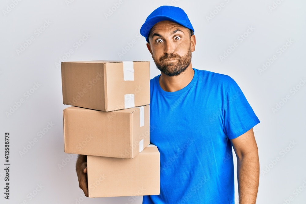 Handsome man with beard wearing courier uniform holding delivery packages puffing cheeks with funny face. mouth inflated with air, crazy expression. Handsome man with beard wearing courier uniform holding delivery packages puffing cheeks with funny face. mouth inflated with air, crazy expression.