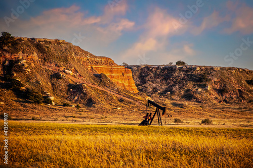 Oil well pump jack in red earth western Oklahoma at golden hour with early sunset sky and bluffs in background.
