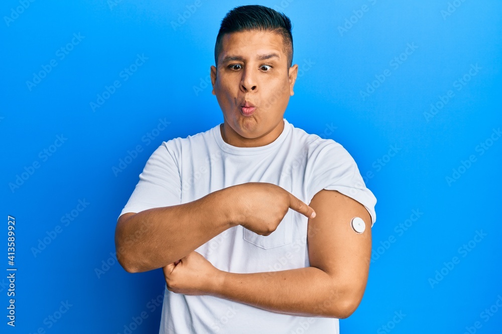Young latin man using glucose meter button on arm making fish face with mouth and squinting eyes, crazy and comical. Young latin man using glucose meter button on arm making fish face with mouth and squinting eyes, crazy and comical.