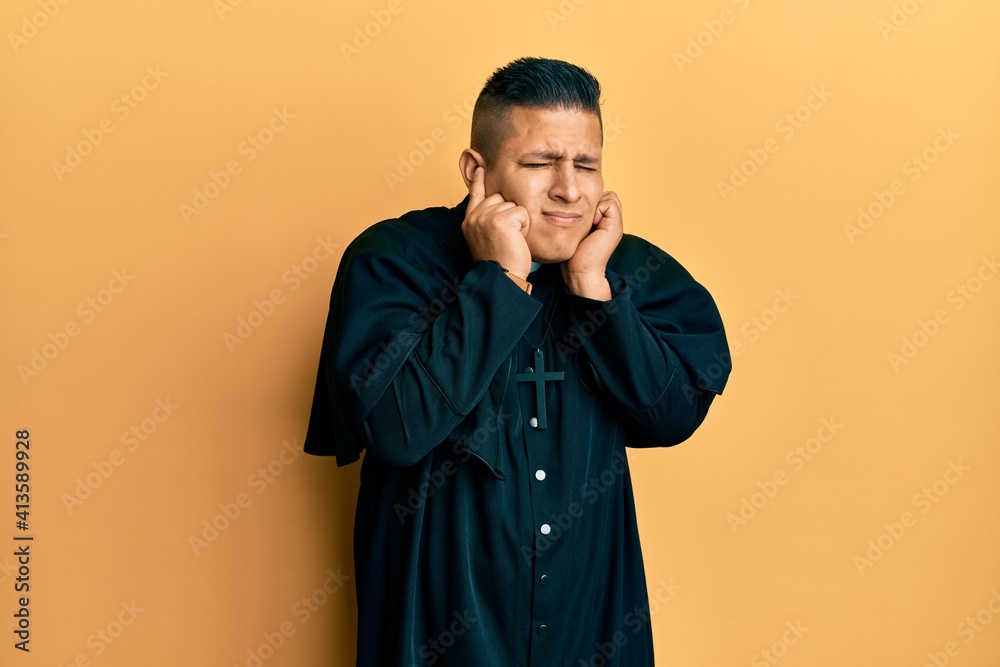 Young latin priest man standing over yellow background covering ears with fingers with annoyed expression for the noise of loud music. deaf concept.
