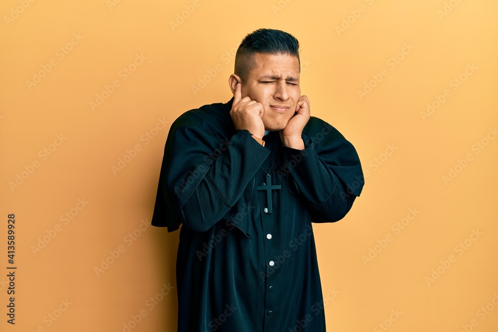 Young latin priest man standing over yellow background covering ears with fingers with annoyed expression for the noise of loud music. deaf concept. Young latin priest man standing over yellow background covering ears with fingers with annoyed expression for the noise of loud music. deaf concept.