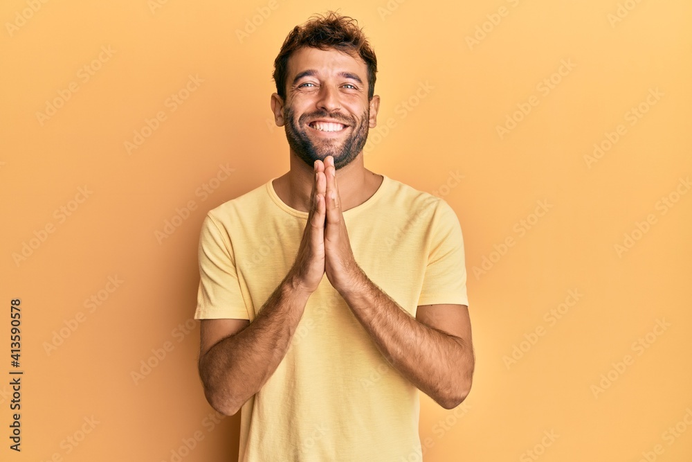 Handsome man with beard wearing casual yellow tshirt over yellow background praying with hands together asking for forgiveness smiling confident.