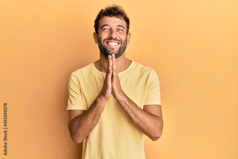 Handsome man with beard wearing casual yellow tshirt over yellow background praying with hands together asking for forgiveness smiling confident. Handsome man with beard wearing casual yellow tshirt over yellow background praying with hands together asking for forgiveness smiling confident.