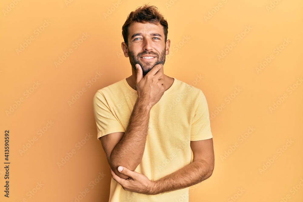 Handsome man with beard wearing casual yellow tshirt over yellow background looking confident at the camera smiling with crossed arms and hand raised on chin. thinking positive. Handsome man with beard wearing casual yellow tshirt over yellow background looking confident at the camera smiling with crossed arms and hand raised on chin. thinking positive.