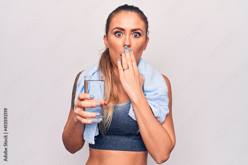 Beautiful blonde sporty woman using towel drinking bottle of water over white background covering mouth with hand, shocked and afraid for mistake. Surprised expression