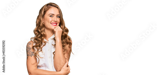 Young blonde girl wearing casual clothes looking confident at the camera with smile with crossed arms and hand raised on chin. thinking positive.