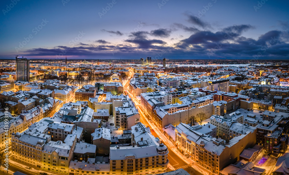 Riga city panorama in winter time. Street lights glowing in blue hour. Snowy rooftops. Iconic buildings and architecture. Drone footage. 