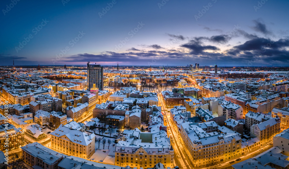 Obraz premium Riga city panorama in winter time. Street lights glowing in blue hour. Snowy rooftops. Iconic buildings and architecture. Drone footage. 