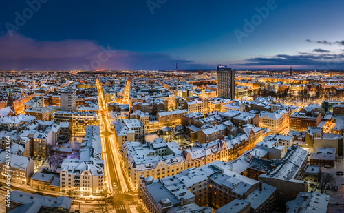 Riga city panorama in winter time. Street lights glowing in blue hour. Snowy rooftops. Iconic buildings and architecture. Drone footage. 