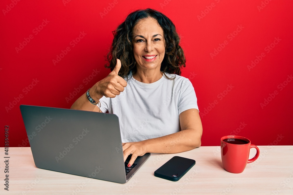 Fototapeta premium Beautiful middle age woman working at the office using computer laptop smiling happy and positive, thumb up doing excellent and approval sign