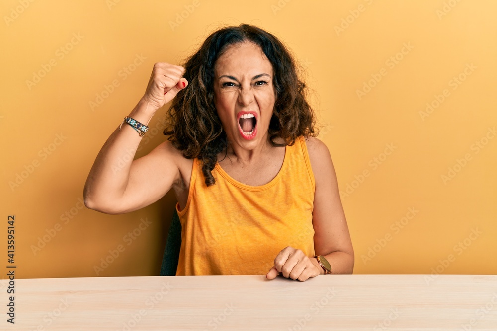 Beautiful middle age woman wearing casual clothes sitting on the table angry and mad raising fist frustrated and furious while shouting with anger. rage and aggressive concept. Beautiful middle age woman wearing casual clothes sitting on the table angry and mad raising fist frustrated and furious while shouting with anger. rage and aggressive concept.