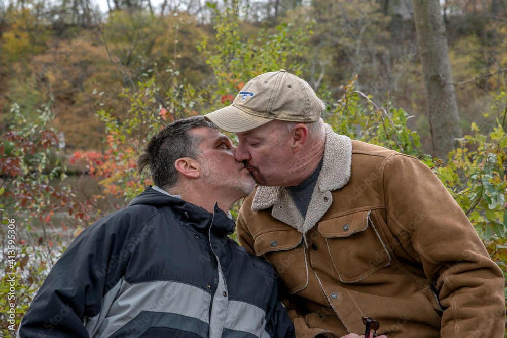 An LGBTQ middle-aged gay couple kissing outdoors on autumn day.