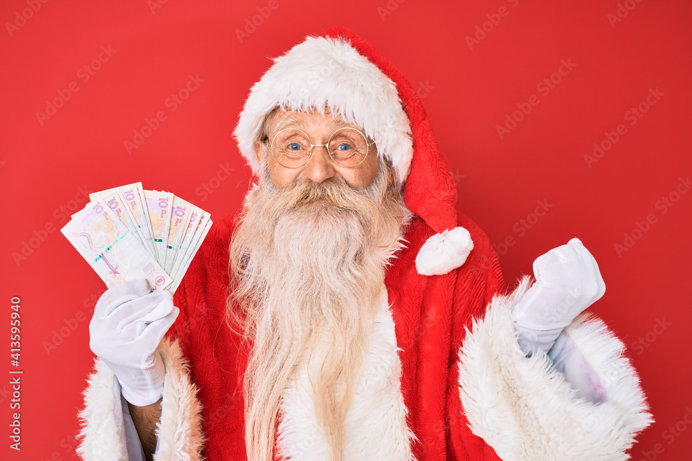 Old senior man with grey hair and long beard wearing santa claus costume holding colombian pesos screaming proud, celebrating victory and success very excited with raised arm Old senior man with grey hair and long beard wearing santa claus costume holding colombian pesos screaming proud, celebrating victory and success very excited with raised arm
