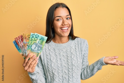 Beautiful hispanic woman holding australian dollars celebrating achievement with happy smile and winner expression with raised hand