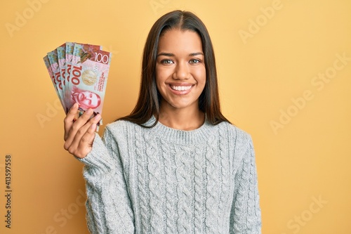 Beautiful hispanic woman holding 100 new zealand dollars banknote looking positive and happy standing and smiling with a confident smile showing teeth