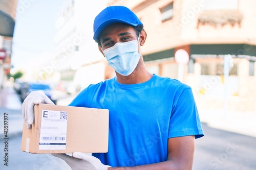 African delivery man wearing courier uniform outdoors wearing coronavirus safety mask holding cardboard parcel © Krakenimages.com
