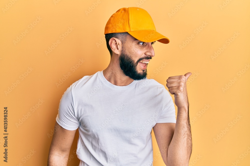 Young man with beard wearing yellow cap pointing thumb up to the side smiling happy with open mouth