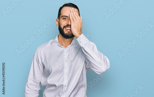 Young man with beard wearing business shirt covering one eye with hand, confident smile on face and surprise emotion.