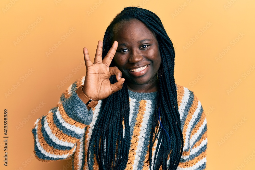 Young black woman with braids wearing casual winter sweater smiling positive doing ok sign with hand and fingers. successful expression. Young black woman with braids wearing casual winter sweater smiling positive doing ok sign with hand and fingers. successful expression.