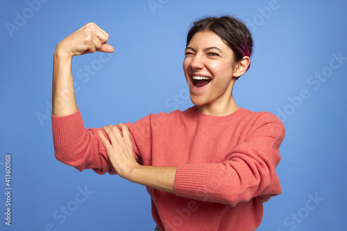 Self assured confident young sporty female raising hand, demonstrating her arm muscles, tensing bicep, feeling strong and independent, opening mouth widely, winking. Strength, sport and femininity