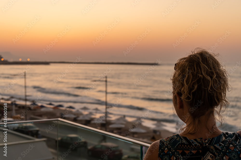 Fototapeta premium Blond girl with curly hair looking towards Old Venetian harbor and beach with umbrellas in the evening in city of Rethymno, Crete, Greece