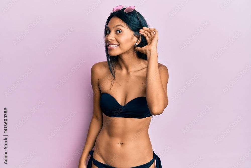 Young african american woman wearing bikini smiling with hand over ear listening and hearing to rumor or gossip. deafness concept. Young african american woman wearing bikini smiling with hand over ear listening and hearing to rumor or gossip. deafness concept.