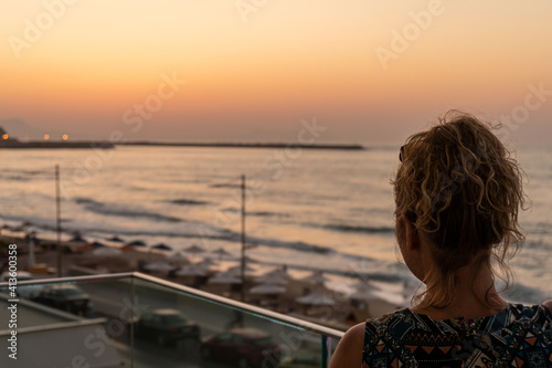 Fototapeta Naklejka Na Ścianę i Meble -  Blond girl with curly hair looking towards Old Venetian harbor and beach with umbrellas in the evening in city of Rethymno, Crete, Greece