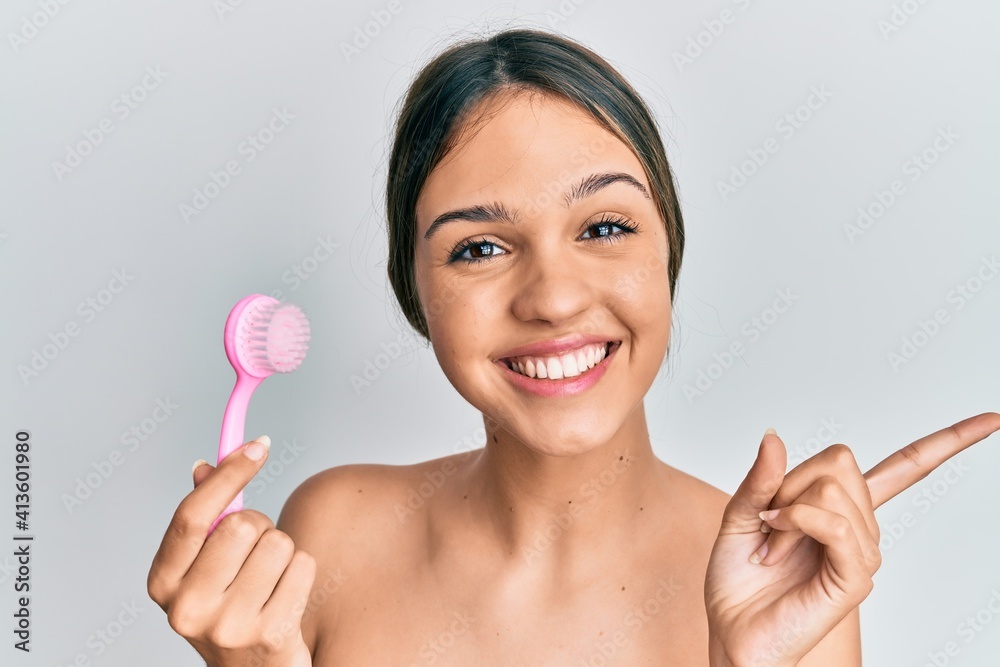 Young brunette woman using facial exfoliating brush smiling happy pointing with hand and finger to the side