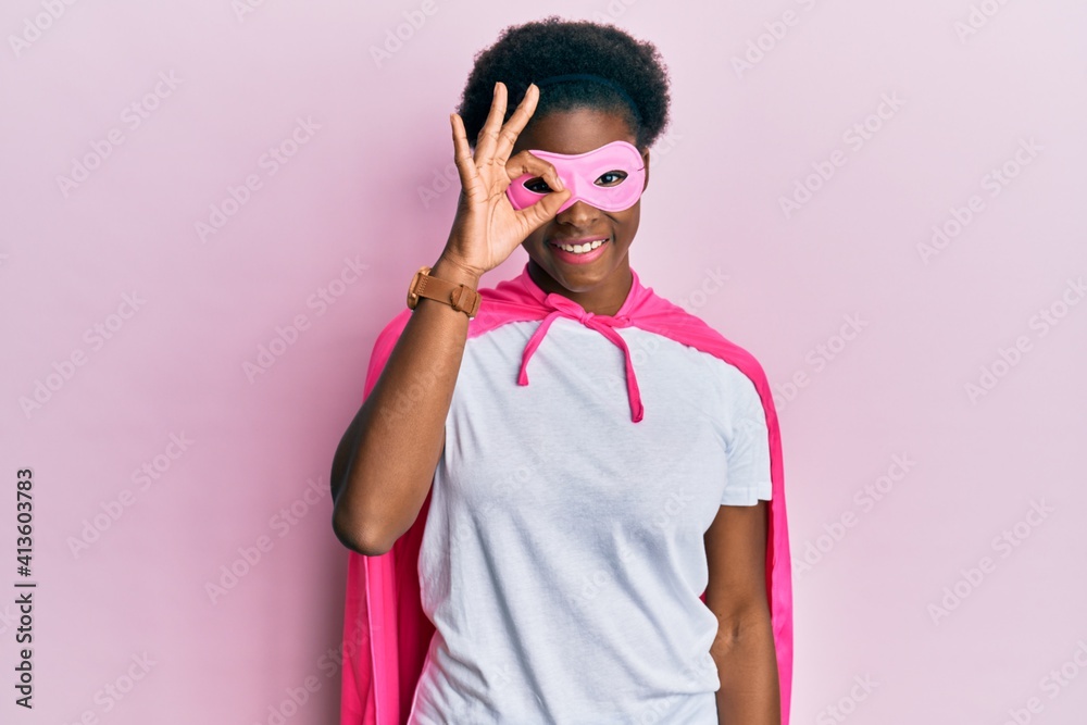 Young african american girl wearing superhero mask and cape costume doing ok gesture with hand smiling, eye looking through fingers with happy face. Young african american girl wearing superhero mask and cape costume doing ok gesture with hand smiling, eye looking through fingers with happy face.