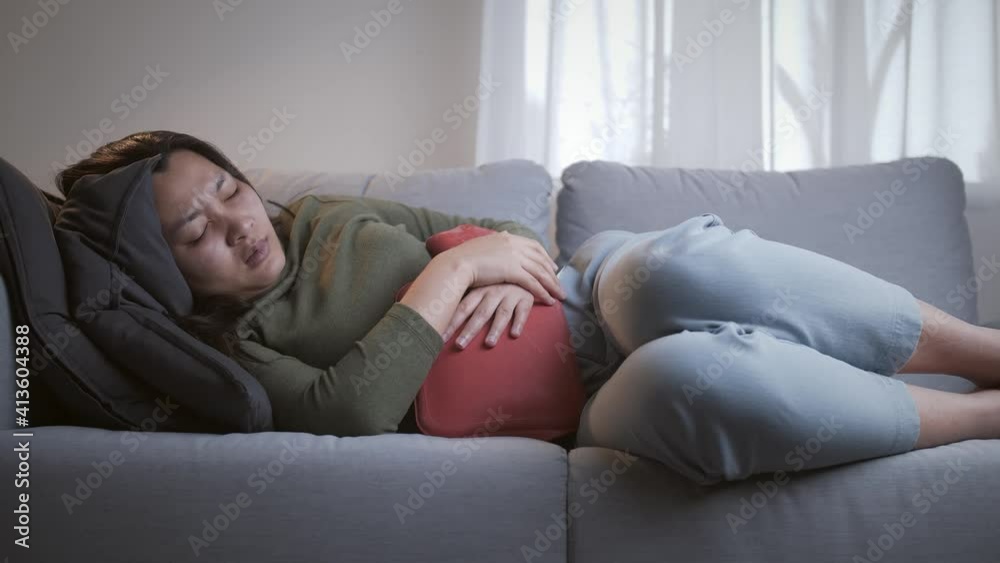 Sick woman with heavy menstrual cramps is applying a hot water bag on her stomach to relieve the pain and feel better on the living room sofa Sick woman with heavy menstrual cramps is applying a hot water bag on her stomach to relieve the pain and feel better on the living room sofa