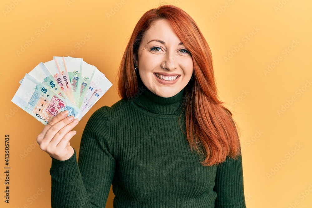 Beautiful redhead woman holding singapore dollars banknotes looking positive and happy standing and smiling with a confident smile showing teeth Beautiful redhead woman holding singapore dollars banknotes looking positive and happy standing and smiling with a confident smile showing teeth