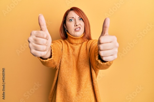 Beautiful redhead woman doing thumbs up positive gesture in shock face, looking skeptical and sarcastic, surprised with open mouth photo