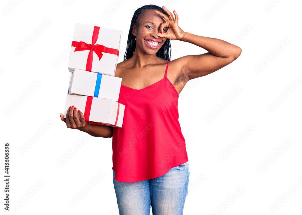 Young african american woman holding gift smiling happy doing ok sign with hand on eye looking through fingers Young african american woman holding gift smiling happy doing ok sign with hand on eye looking through fingers